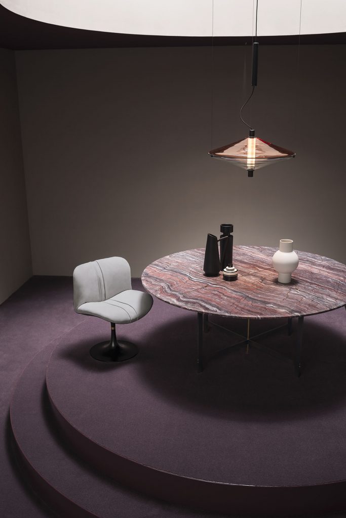 A Placè Dining Table. Base in brass with bronzing oxidation on the flat part and satin finish on the thickness, metal base varnished with burnished effect and satin brass details. Leg connection plate in matte black metal. Rocky mountain stone top with water-repellent protective oil with reinforcing aluminium honeycomb support. Decorative inserts in satin-finished brass with wax finish in a dining room.