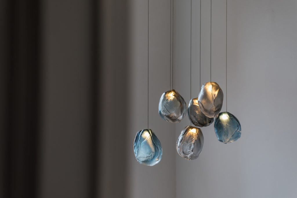 A Seventy Three V lamp in black and blue blown glass in fabric shape, braided metal coaxial cable, electrical components, brushed nickel in a grey background.