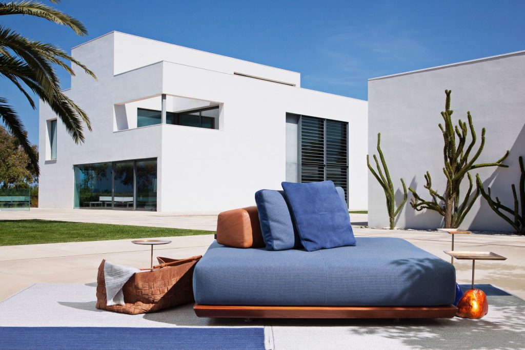 Caprera Island sofa. Upholstered in blue fabric, base in brown leather and blue and brown cushions on a terrace.