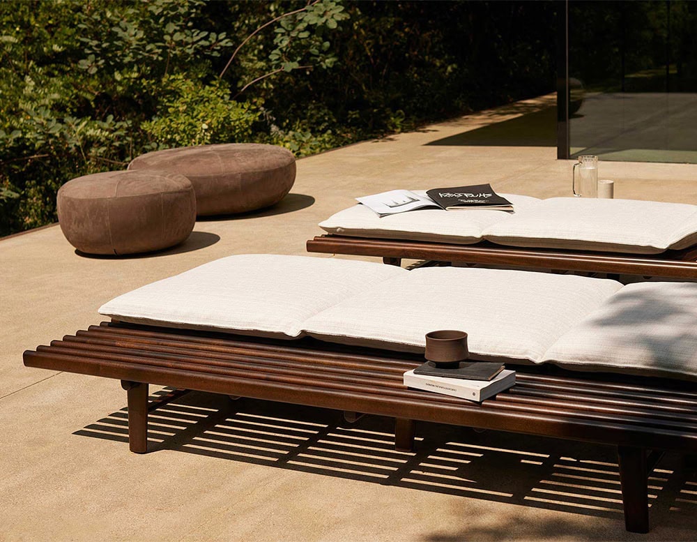 Two 10th Clove Outdoor Poufs are placed on a patio near lounge chairs with white cushions and dark wood frames, under a sunny sky