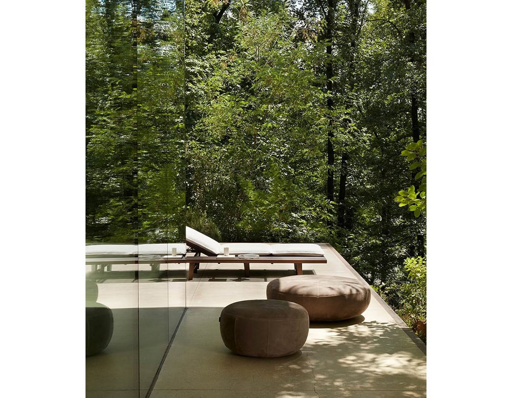 Two 10th Clove Outdoor Poufs are positioned on a patio overlooking a dense green forest, with a lounge chair visible in the background
