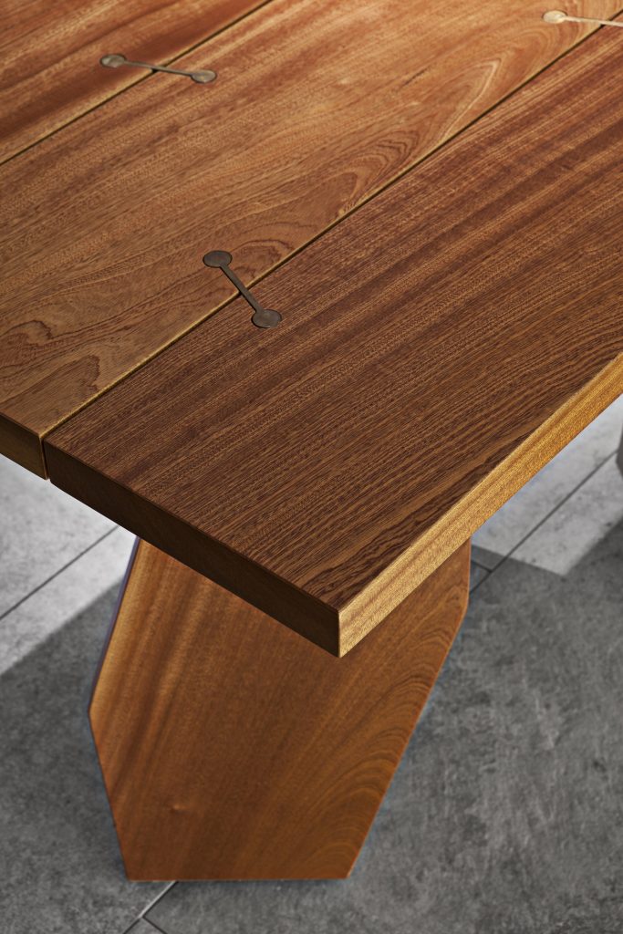 Closeup of 10th Joint Dinner Table: Long rectangular top in brown wood materic slabs connected by burnished brass joints and two polygonal legs on a gray background.