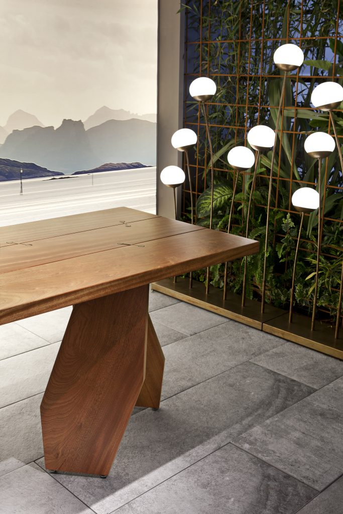Closeup of 10th Joint Dinner Table: Long rectangular top in brown wood materic slabs connected by burnished brass joints and two polygonal legs on a gray background.