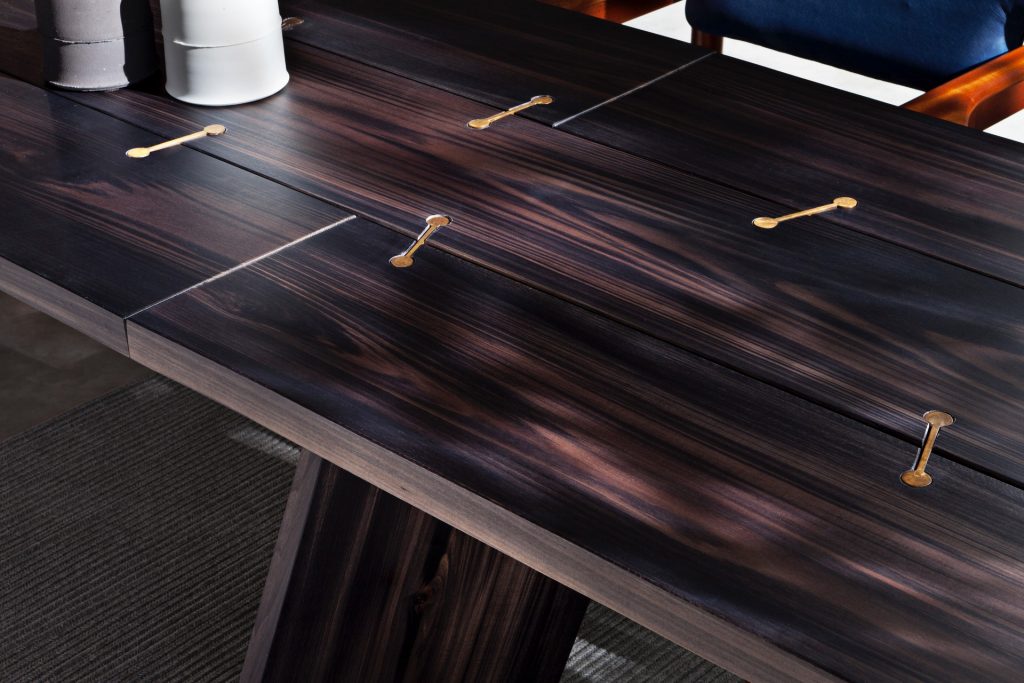 Closeup of 10th Joint Dinner Table: Long rectangular top in brown wood materic slabs connected by burnished brass joints and two polygonal legs on a gray background.