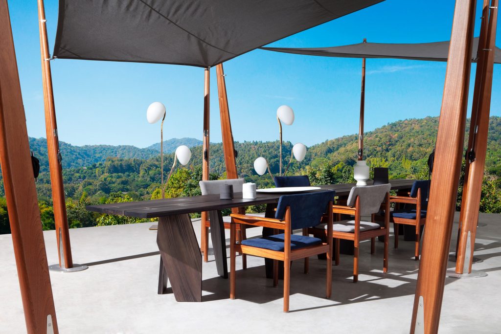 10th Joint Dinner Table: Long rectangular top in brown wood materic slabs connected by burnished brass joints and two polygonal legs on a terrace.