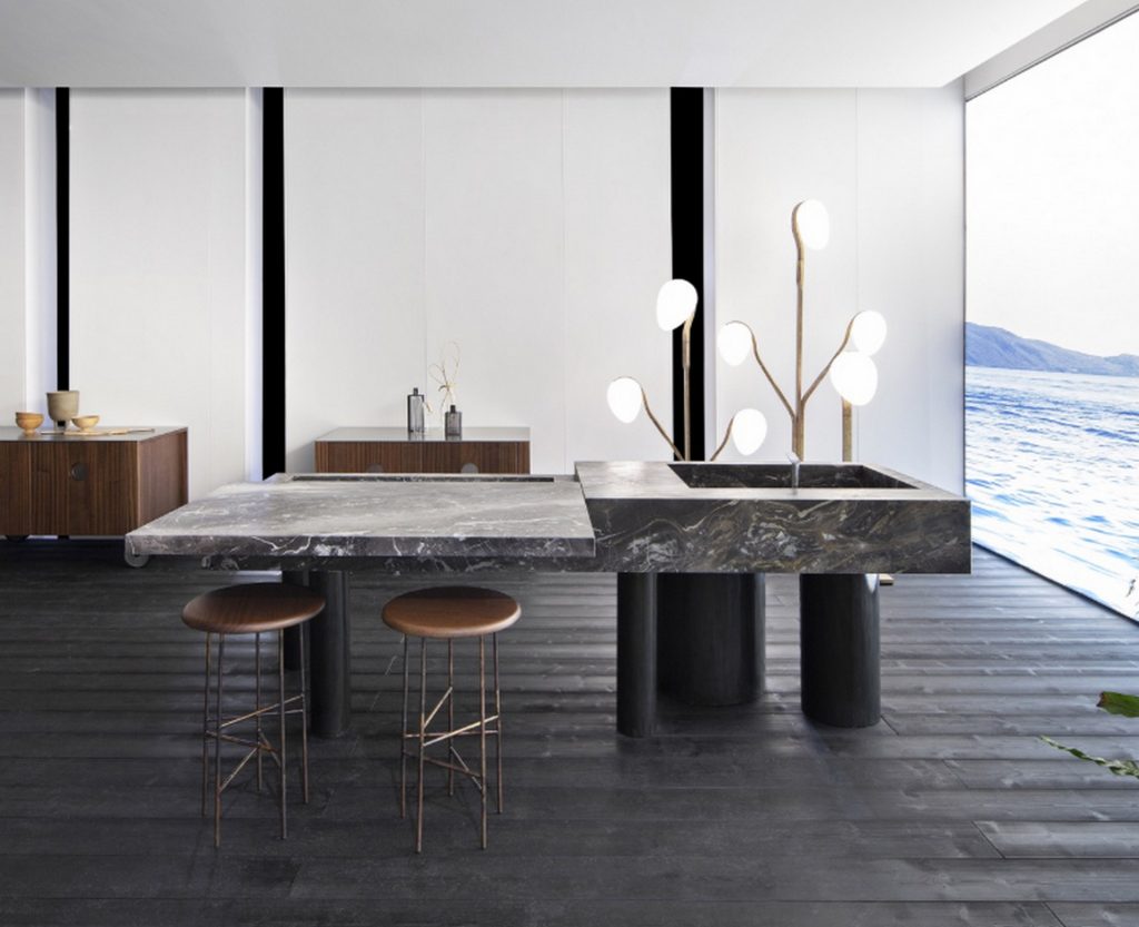 10th Kitchen: Modern kitchen setup made of gray stone, a central island and three brass cylindrical legs in a kitchen.