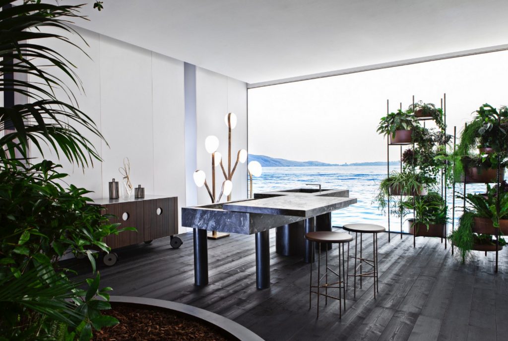 10th Kitchen: Modern kitchen setup made of gray stone, a central island and three brass cylindrical legs in a kitchen.