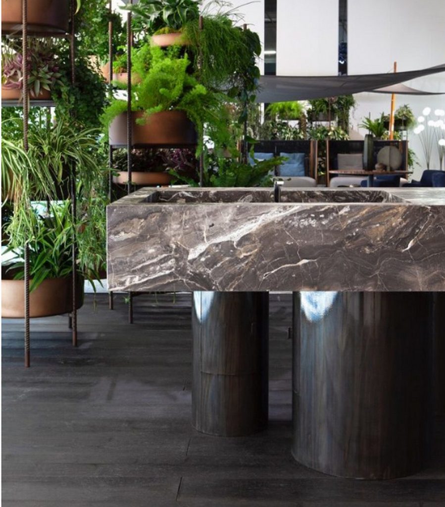 10th Kitchen: Modern kitchen setup made of gray stone, a central island and three brass cylindrical legs in a kitchen.