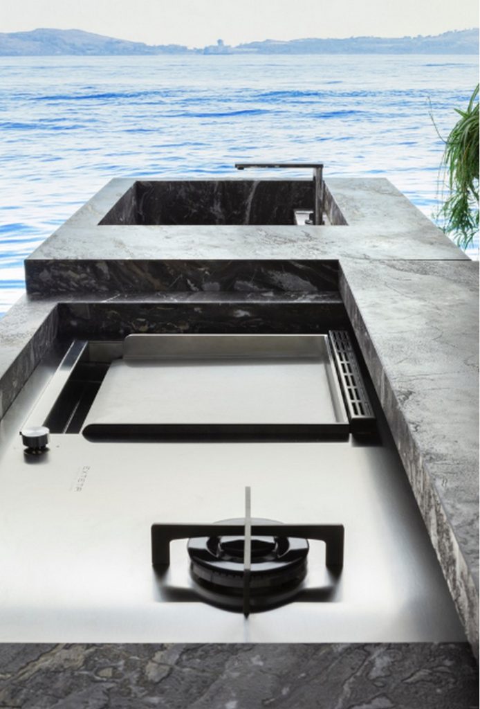 10th Kitchen: Modern kitchen setup made of gray stone, a central island and three brass cylindrical legs in a kitchen.