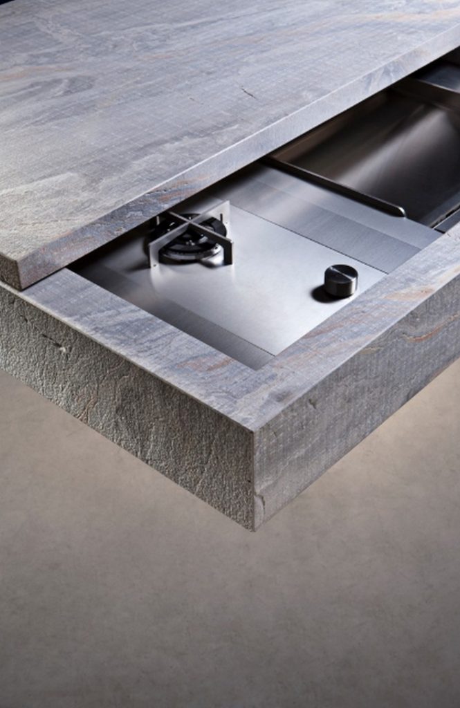 10th Kitchen: Modern kitchen setup made of gray stone, a central island and three brass cylindrical legs in a kitchen.
