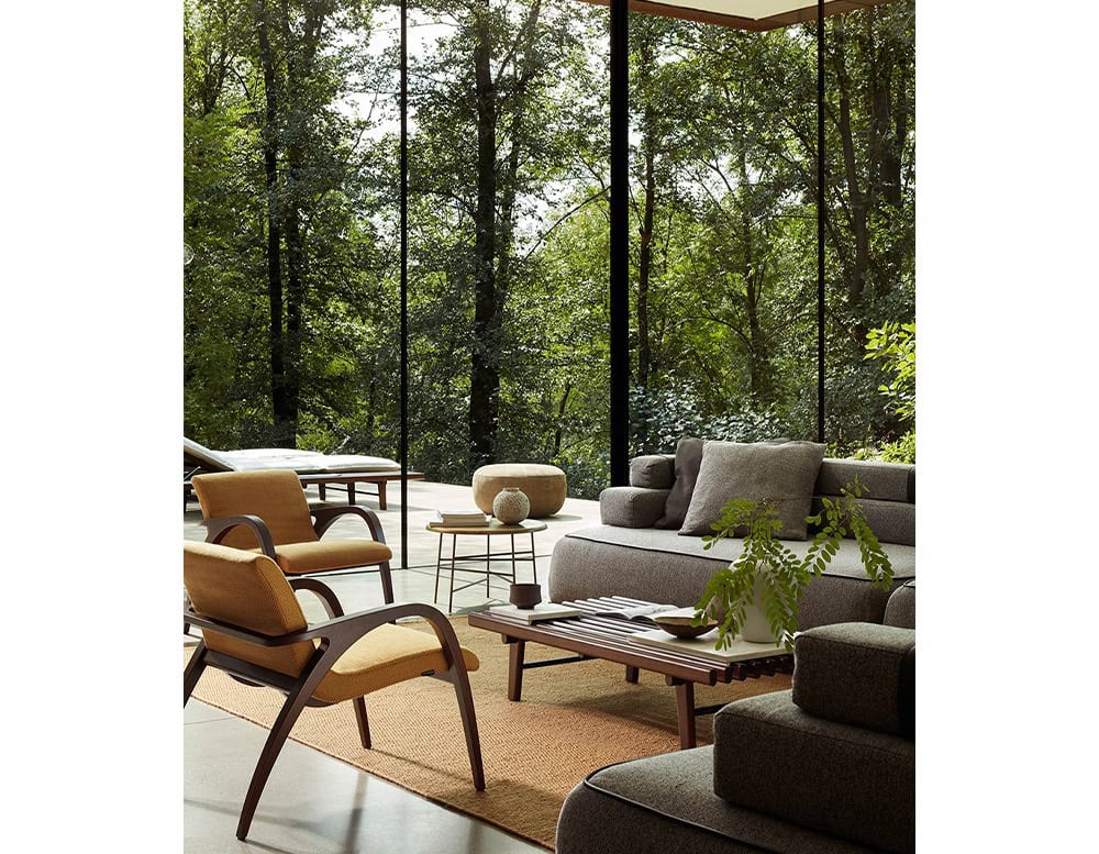 A 10th Star Outdoor Coffee Table Stool is seen among living room furniture including a gray sofa and wooden chairs, with large windows overlooking trees