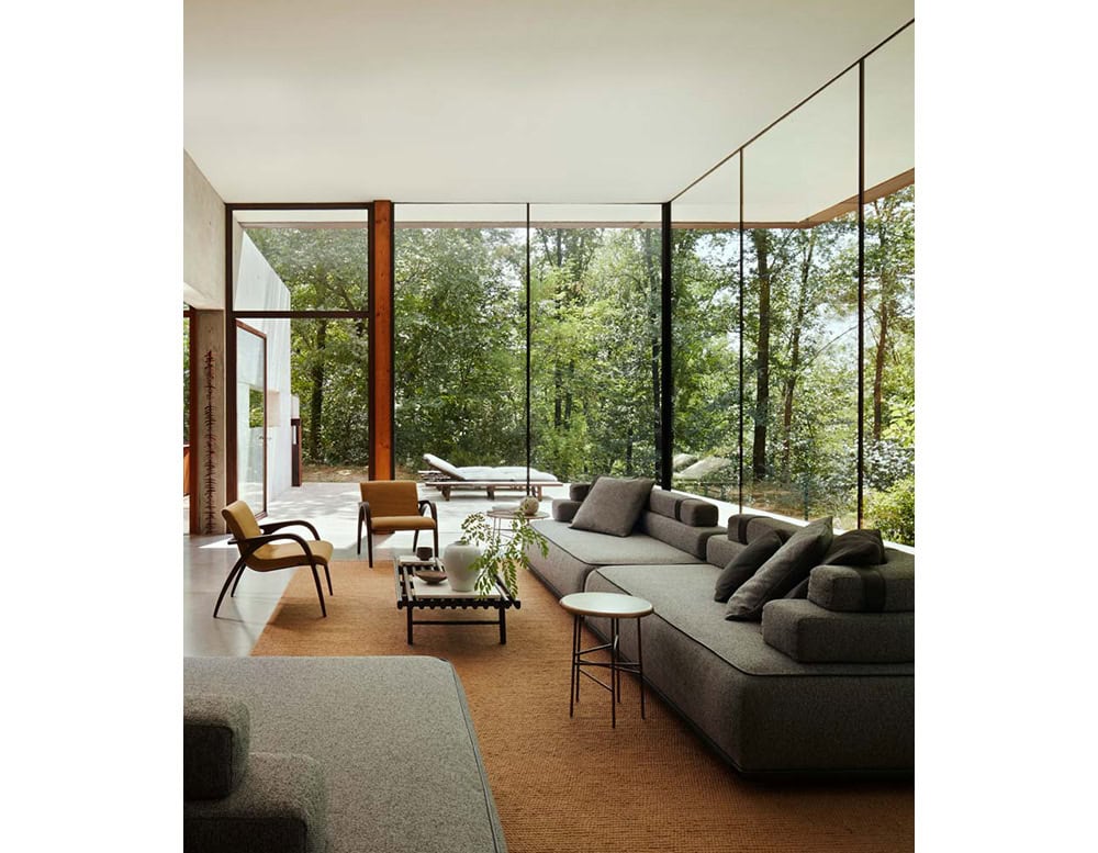 A 10th Star Outdoor Coffee Table Stool with a round top and metal frame is shown next to a sofa and wooden chairs in a living room with a view of trees