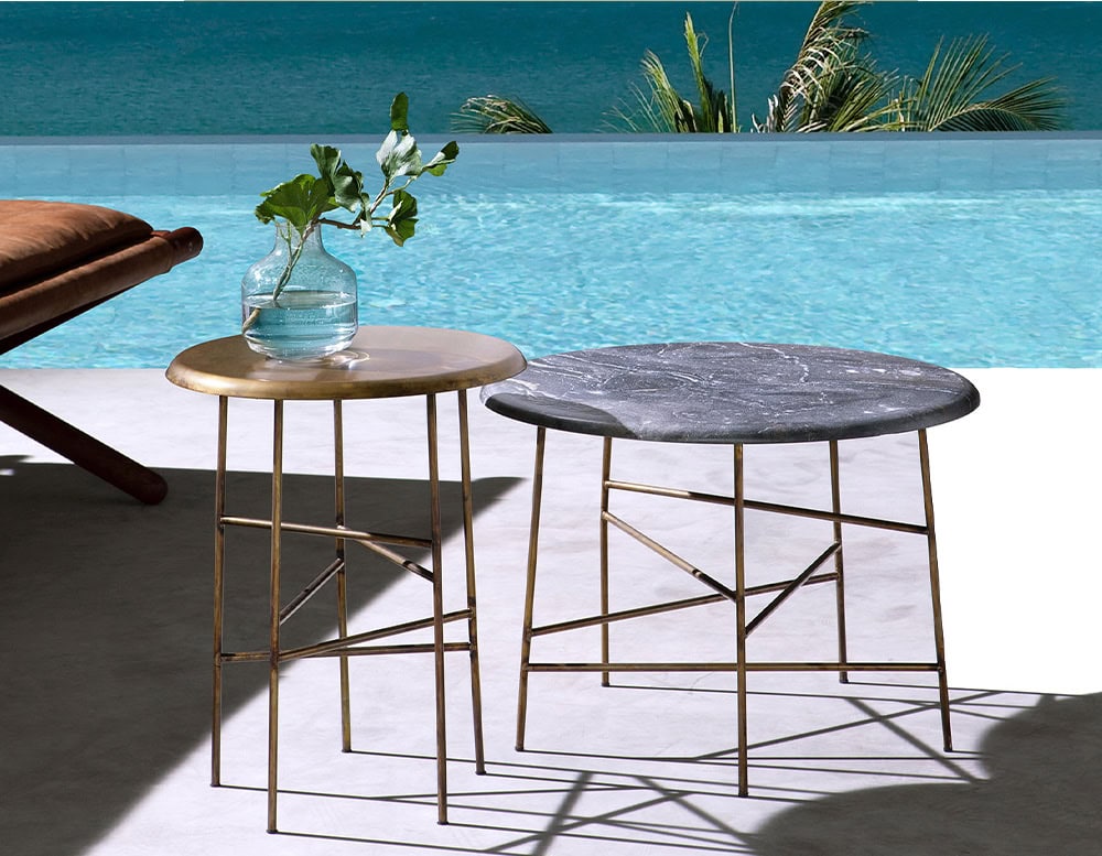 Two 10th Star Outdoor Coffee Table Stools with round tops and metal frames are positioned on a patio beside a pool with a bright blue background