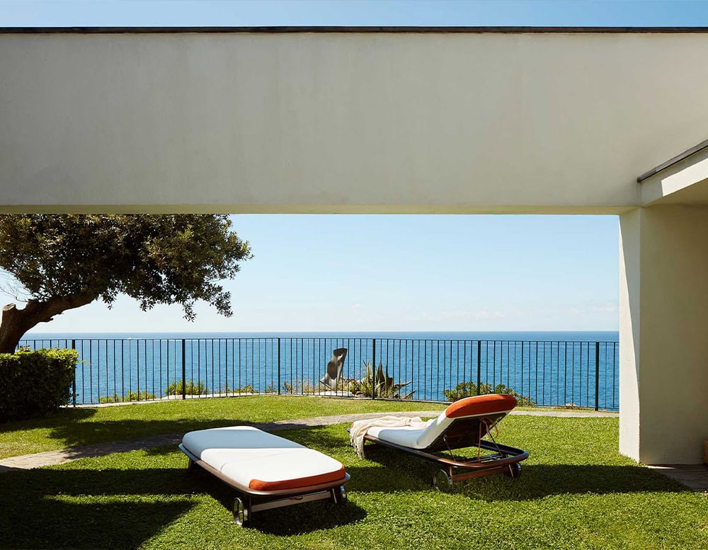 A serene outdoor setting featuring the 10th Tellaro Sun Lounger with plush cushions wooden frames and a breathtaking ocean view under a modern shaded patio
