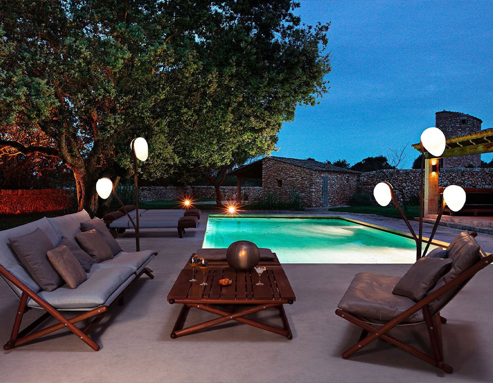 Several 10th Tree Light Outdoor Floor Lights with white, orb-shaped diffusers illuminate a patio with lounge furniture and a swimming pool at dusk