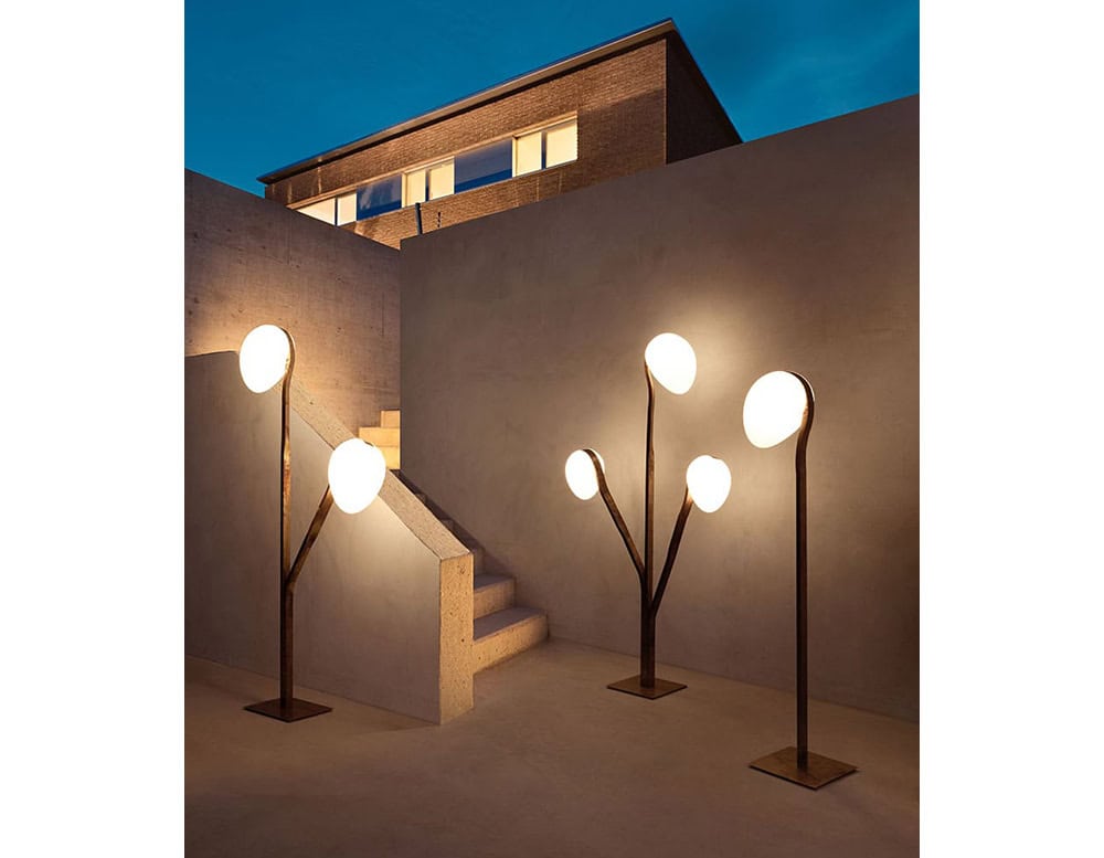 Three 10th Tree Light Outdoor Floor Lights with white, orb-shaped diffusers on slender stems illuminate a patio at dusk, with a building in the background