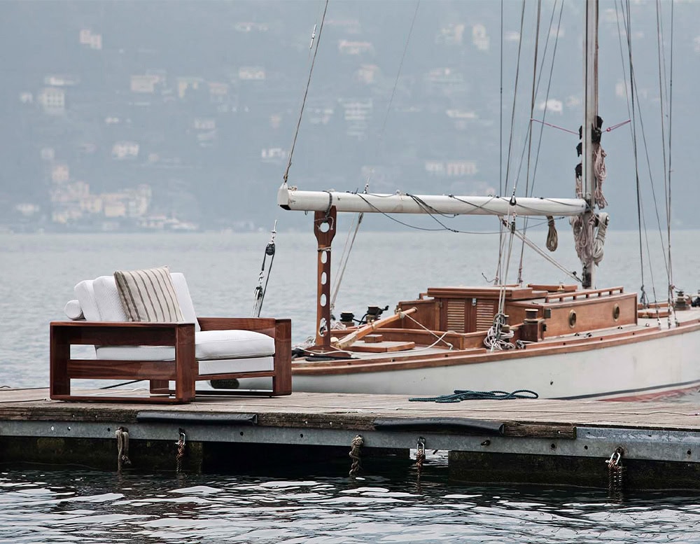 The Bellagio Outdoor Armchair set on a wooden dock featuring a solid wooden frame plush cushions and a scenic lakeside with a sailboat nearby