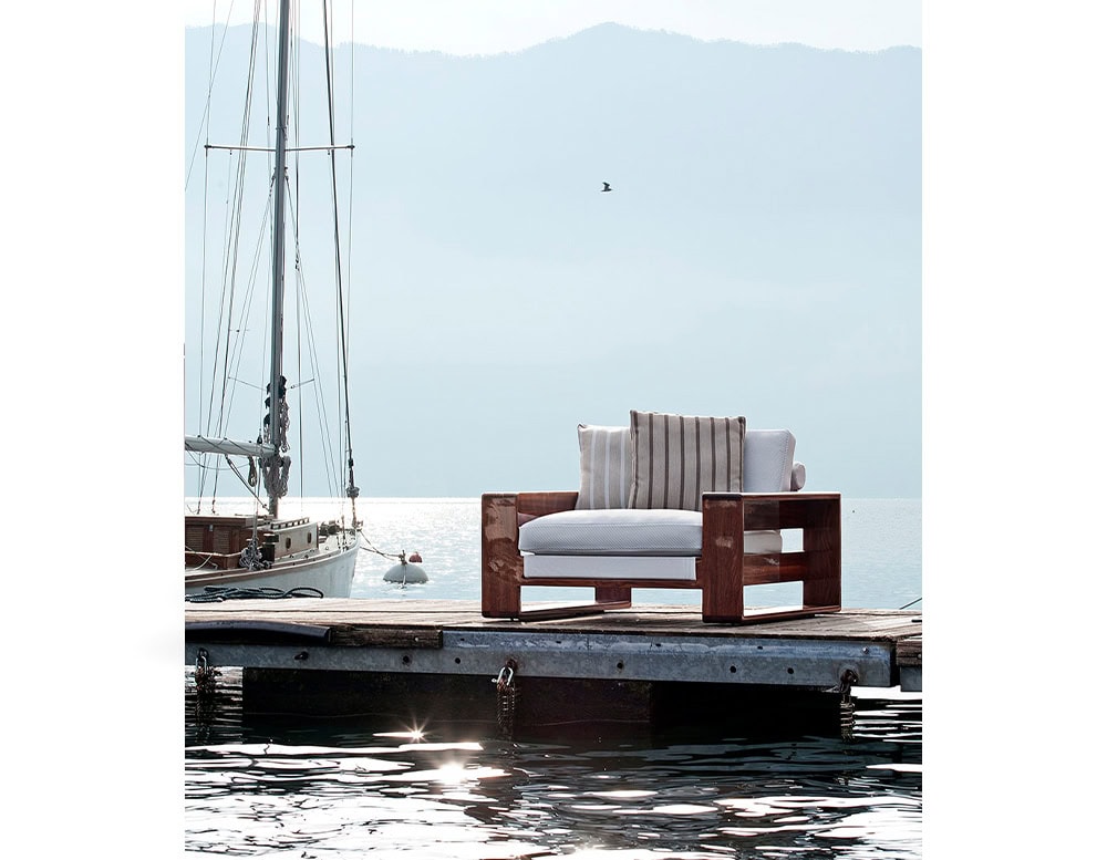 The Bellagio Outdoor Armchair placed on a tranquil dock featuring a solid wooden frame plush cushions and a scenic lakefront with a sailboat