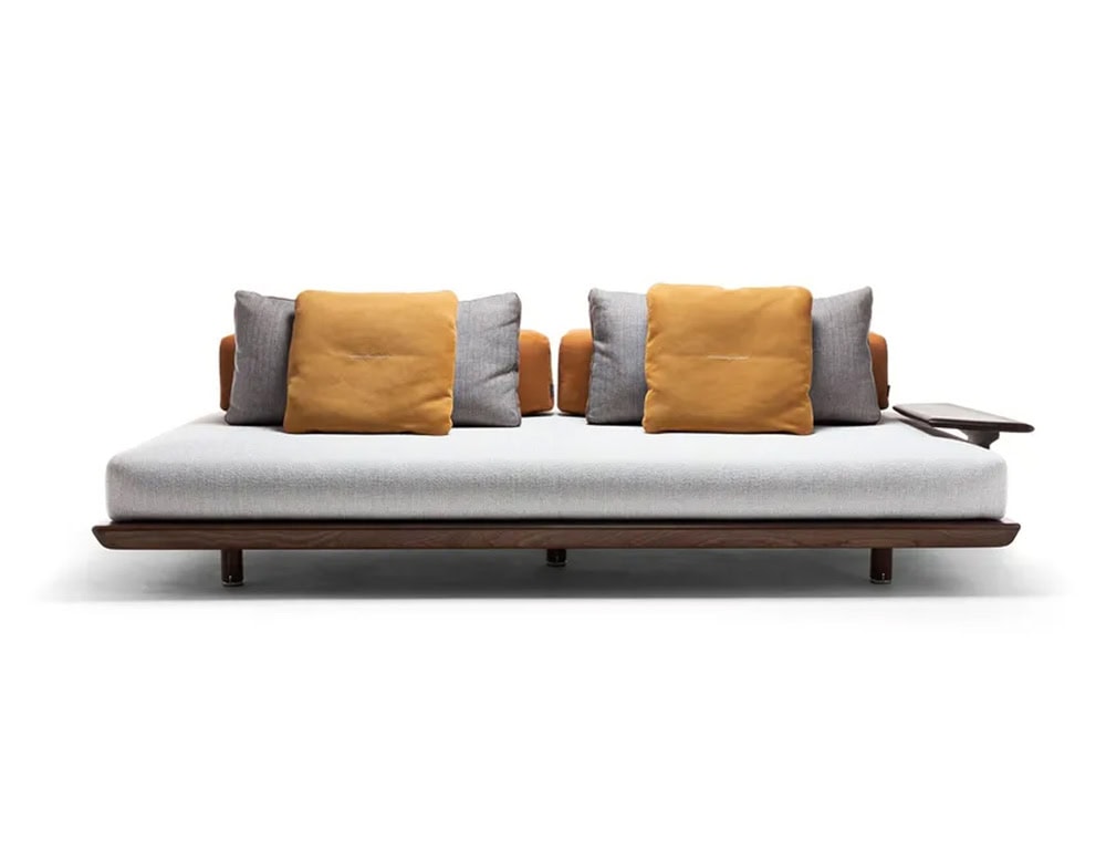 A Caprera Sofa with a light-colored cushion and a dark wood frame is shown against a white background; the sofa has four back cushions, two of which are a contrasting orange color