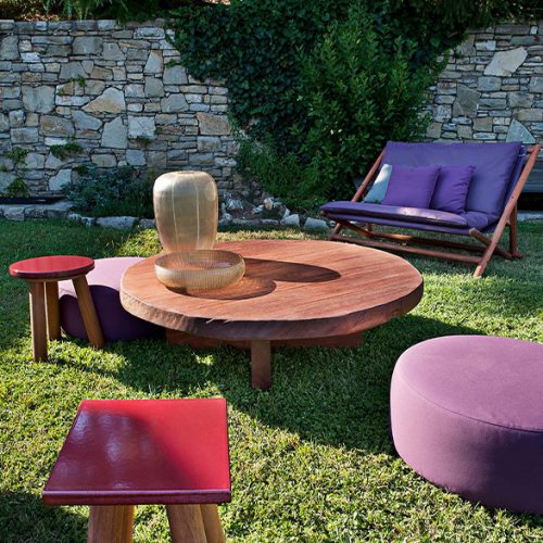 An inviting outdoor lounge featuring the Individual Mini Round table surrounded by colorful seating lush greenery and a stone wall backdrop