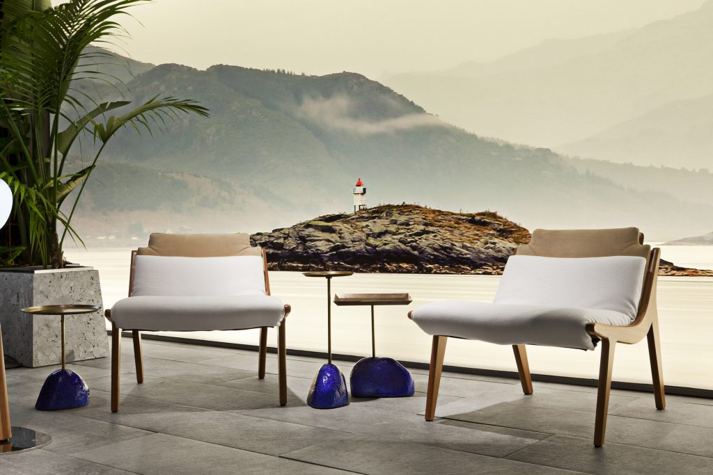Two brown Lerici Armchairs with a curved backrest and thin legs with white cushions on a terrace.
