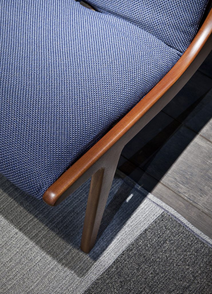 Closeup of brown Lerici Armchair with a curved backrest and thin legs with blue cushions on a gray background.