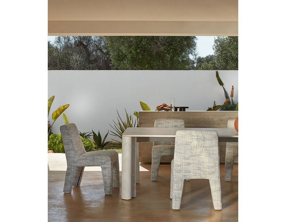 A Michaelangelo Dinner Table and matching chairs are arranged on a patio with a view of greenery and a white wall in the background