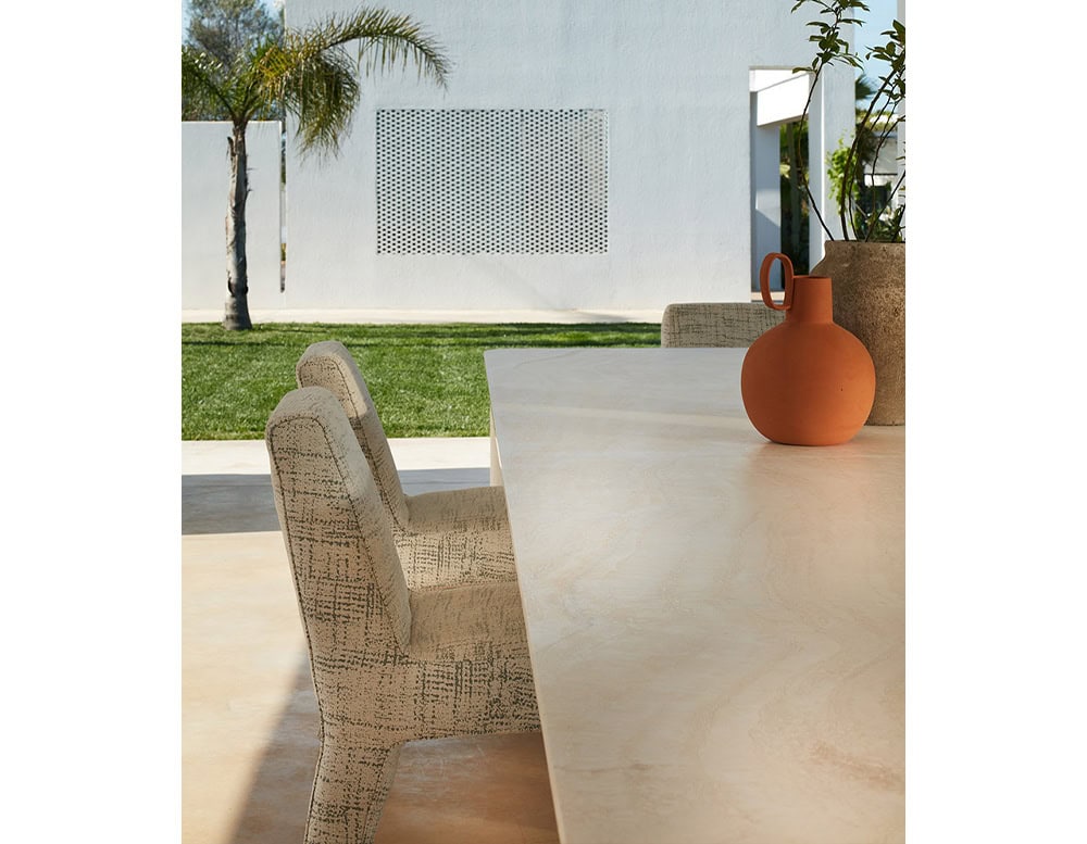 A Michaelangelo Dinner Table with a light-colored surface is partially visible on a patio with a chair and an orange vase, under a sunny sky