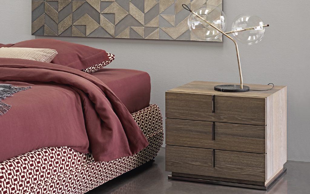 A brown wood Ari Night Table with handles and three drawers in a bedroom.