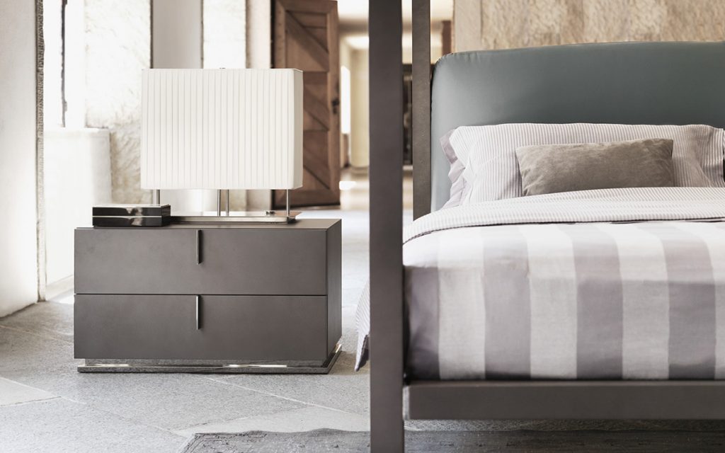 A gray wood Ari Night Table with three drawers and three bronze handles in a bedroom.