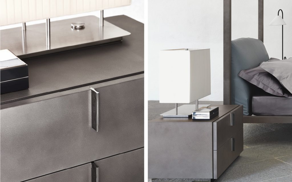 Closeup of a gray wood Ari Night Table with three drawers and handles in a bedroom.