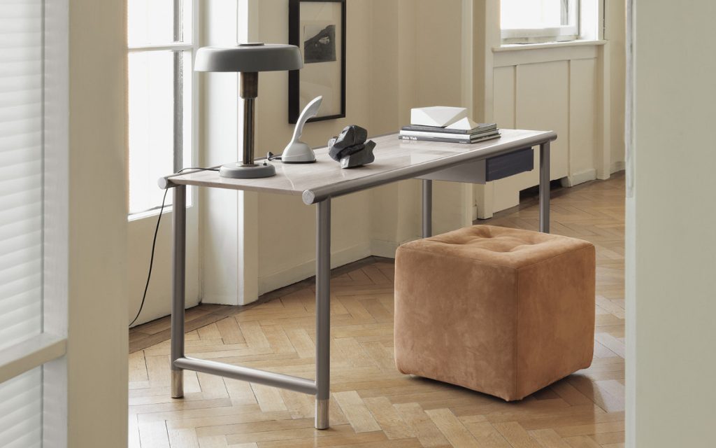 Iko Pouf with cubic shape upholsted in brown fabric in front of a desk.