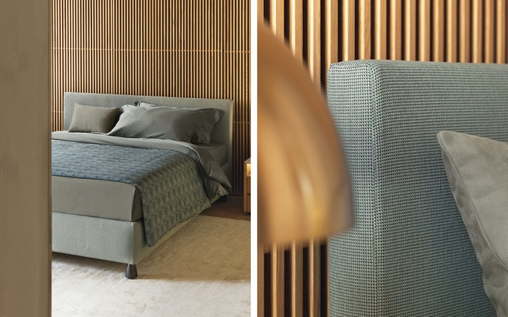 Closeup of Notturno 2 Double bed. Base and backrest covered in gray fabric and bedding in a bedroom.