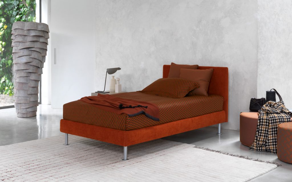 Notturno Single bed. Base and backrest covered in red fabric and legs with bedding in a bedroom.