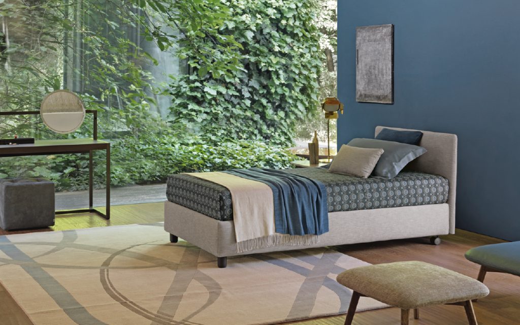 Notturno Single bed. Base and backrest covered in gray fabric and legs with bedding in a bedroom.