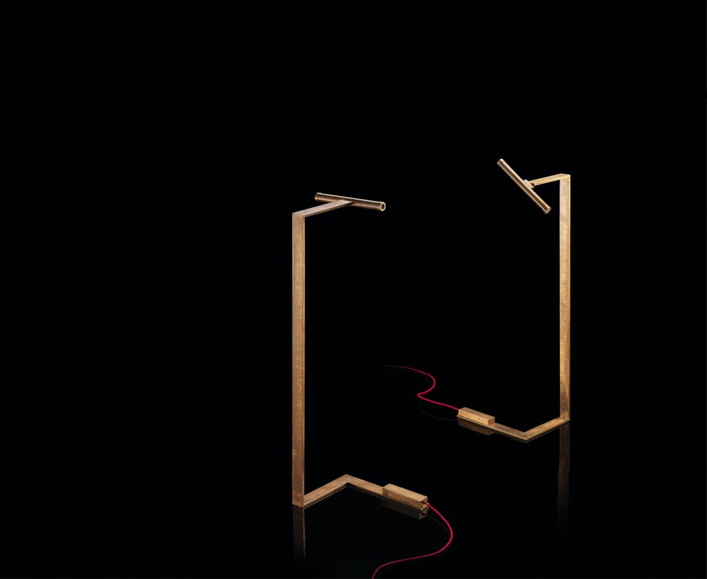 Two Let Flat Lamps with brass pipe structure, multi directional light, power lead with red silk cover, counterweight in stone on a black background.