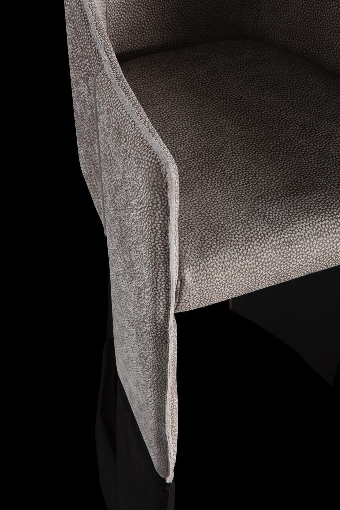 Closeup of Ketch Chair back leg in brass finish. finish padded seat, back, two legs and arms in gray leather finish, back cushion in gray fabric on a black background.