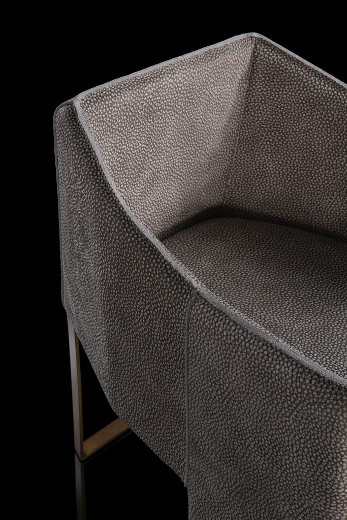 Closeup of Ketch Chair back leg in brass finish. finish padded seat, back, two legs and arms in gray leather finish, back cushion in gray fabric on a black background.