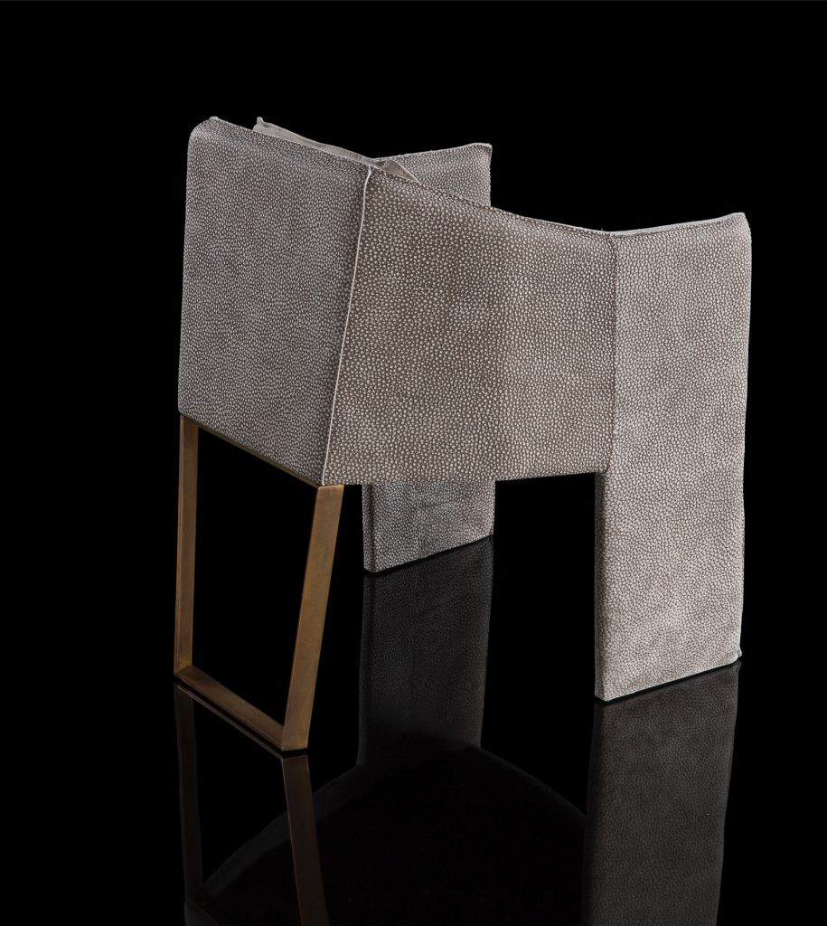 Ketch Chair back leg in brass finish. finish padded seat, back, two legs and arms in gray leather finish, back cushion in gray fabric on a black background.
