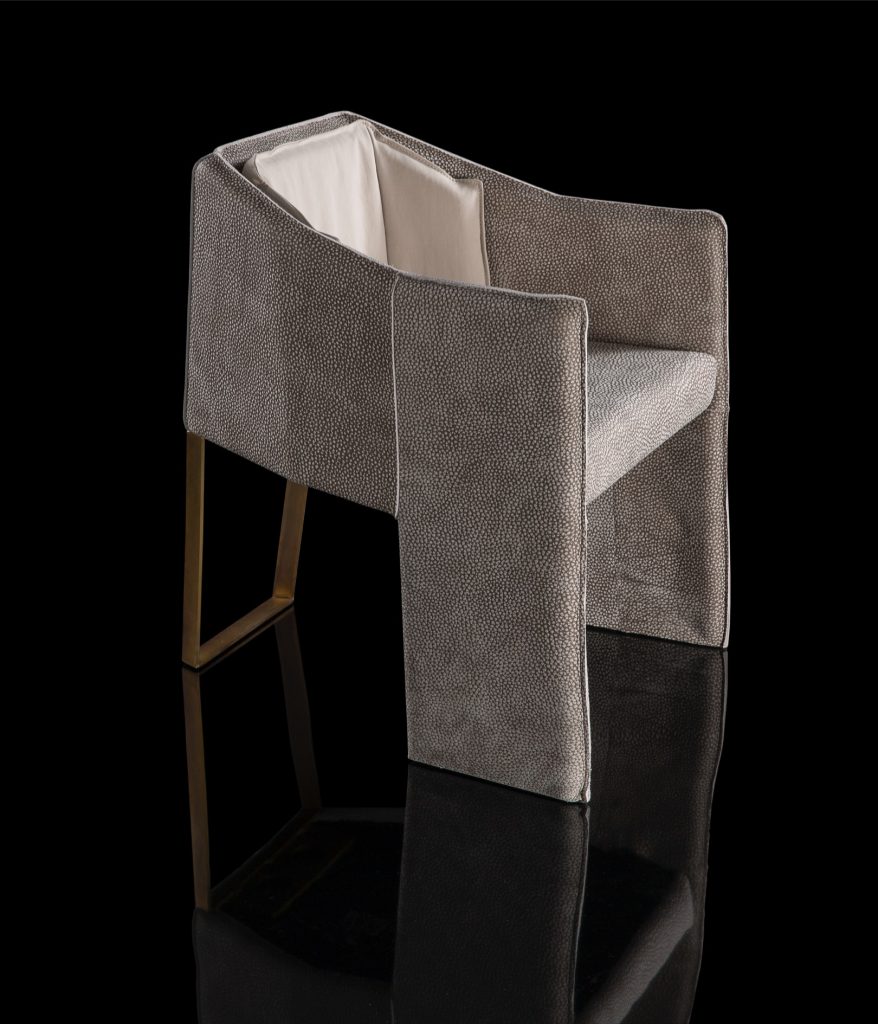Ketch Chair back leg in brass finish. finish padded seat, back, two legs and arms in gray leather finish, back cushion in gray fabric on a black background.