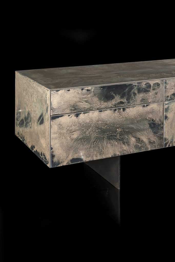 Closeup of Noctis Vanity cabinet with three doors. Outside covered with burnished steel, three legs in black steel on a black background.