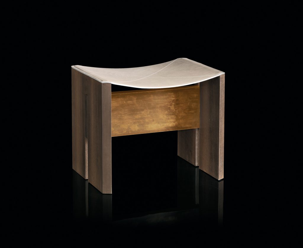 Rio Stool, structure and two legs in natural wood with central crosspiece covered in brass; seat in gray leather on a black background.
