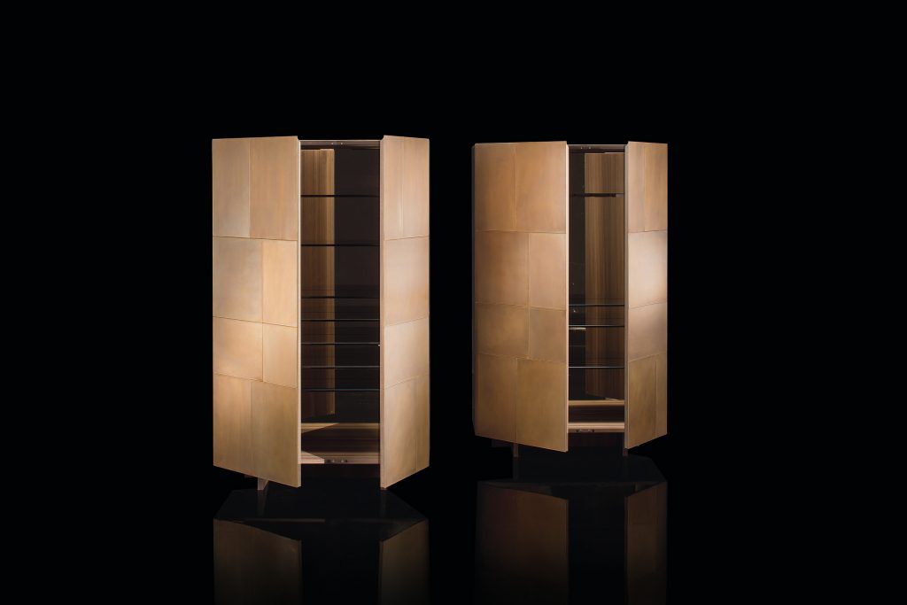 Two Slim Sides Vertical, doors finish and two legs in brass with a pattern of squares and rectangles, back in bronzed mirror on a gray room.