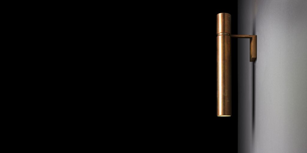 Ceiling Tubular Light with a cylindrical shape in bronze attached to a black wall with a bronze support.