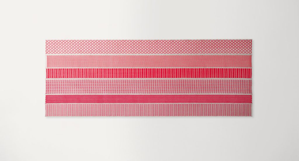 Grafismi modular rug. The geometric pattern is embroidered with red and brown flat braids in Rope yarn on kimia fabric on a white background.