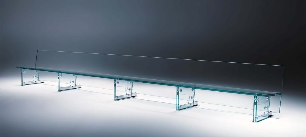 A minimalist glass bench with a sleek, transparent design, featuring a long tempered glass seat supported by two sturdy glass legs against a dark backdrop