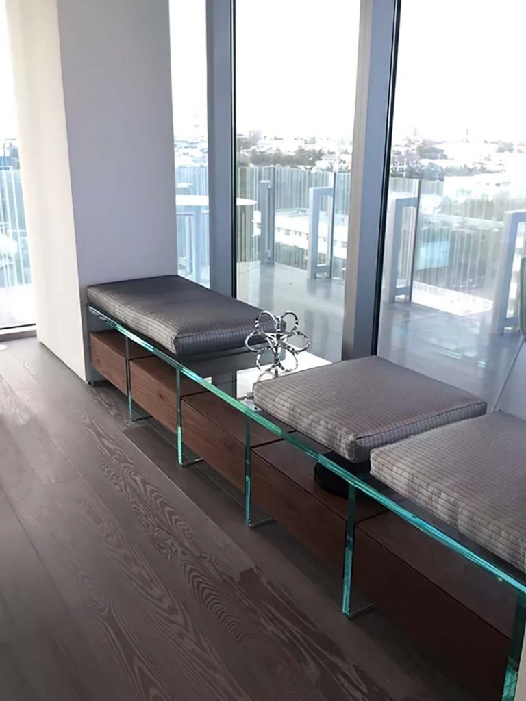 A contemporary glass bench with cushioned seating and integrated wooden storage drawers, positioned along a windowed balcony with a cityscape view