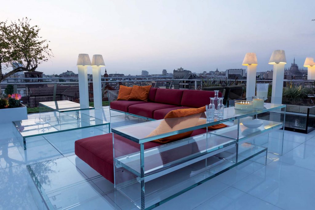 A rooftop terrace at sunset featuring the Simplicity Sofa in red with glass tables and shelves, illuminated by elegant lamps, overlooking the city skyline