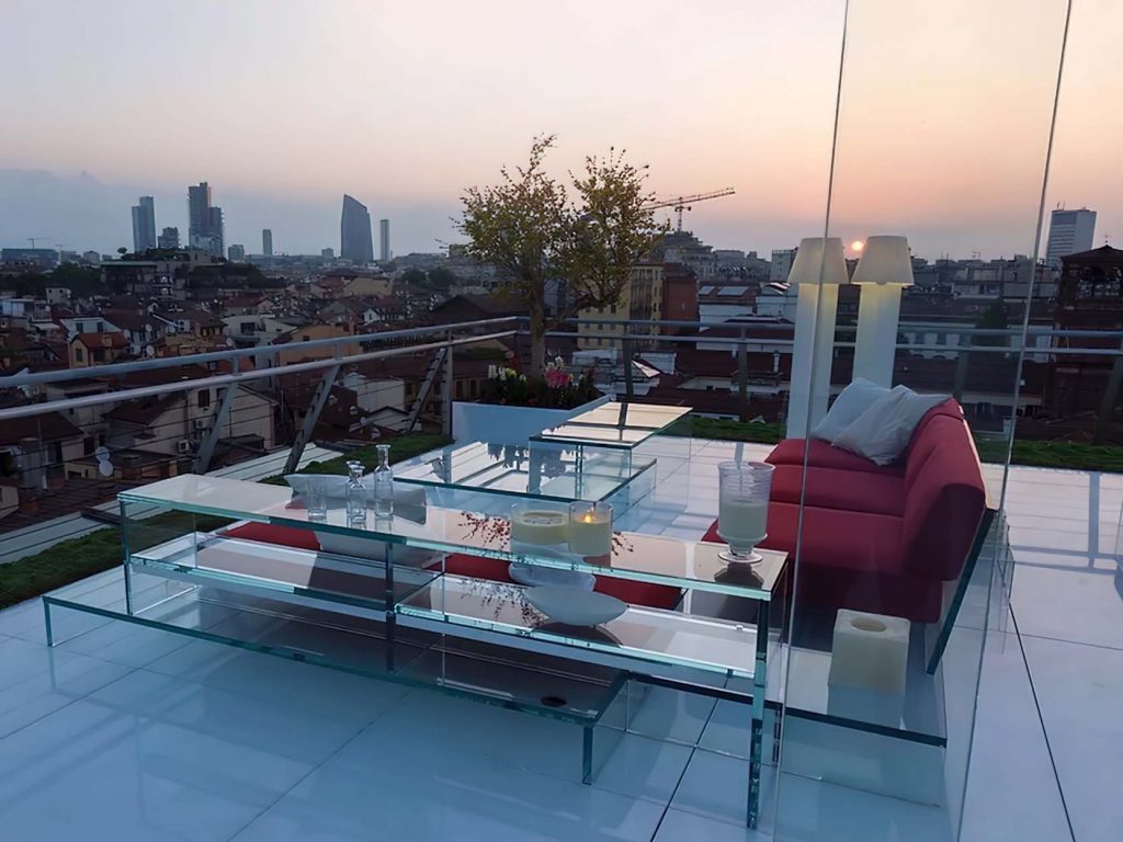 A rooftop terrace at sunset featuring a transparent glass table and shelving unit, a red sofa, and a city skyline in the background