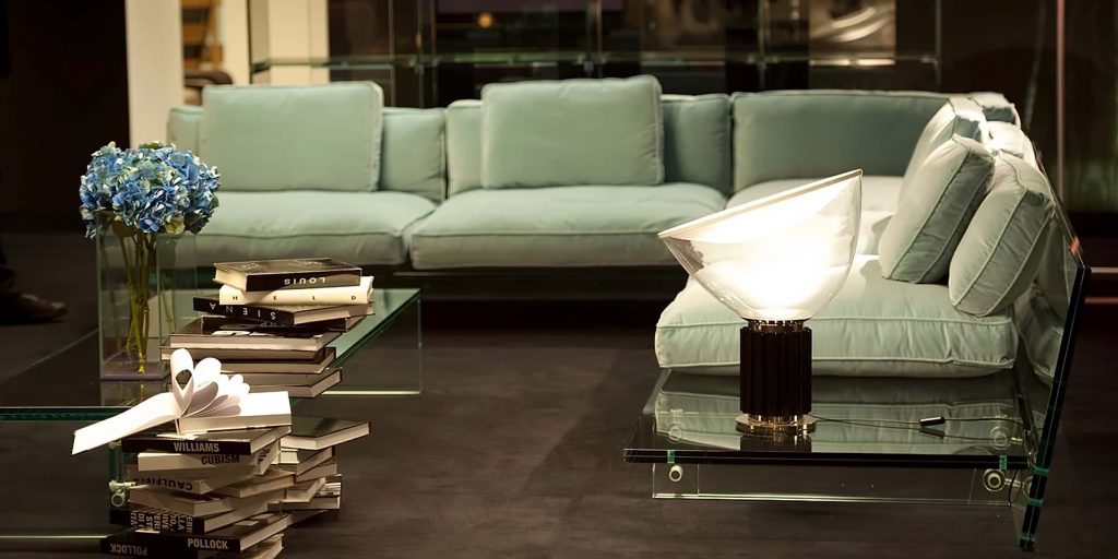 A modern living room featuring a sleek light blue sofa with a transparent glass base, accompanied by a glass coffee table and a sculptural table lamp